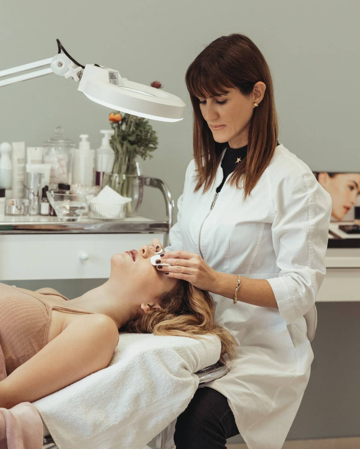 Une esthéticienne en blouse blanche pose des cotons sur les yeux d'une cliente allongée, sous une lampe loupe.